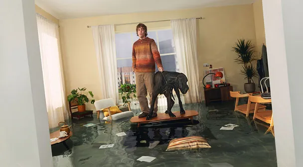 Eine Mann und ein Hund stehen während einer Überschwemmung in einem Wohnraum auf dem Tisch und werden von Wasser umschlossen.